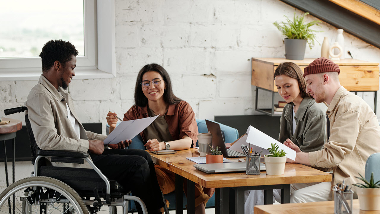 A group of four colleagues are sitting around a table in a bright modern workplace. A young African man is sitting in a wheelchair and is discussing a document with a Hispanic woman.