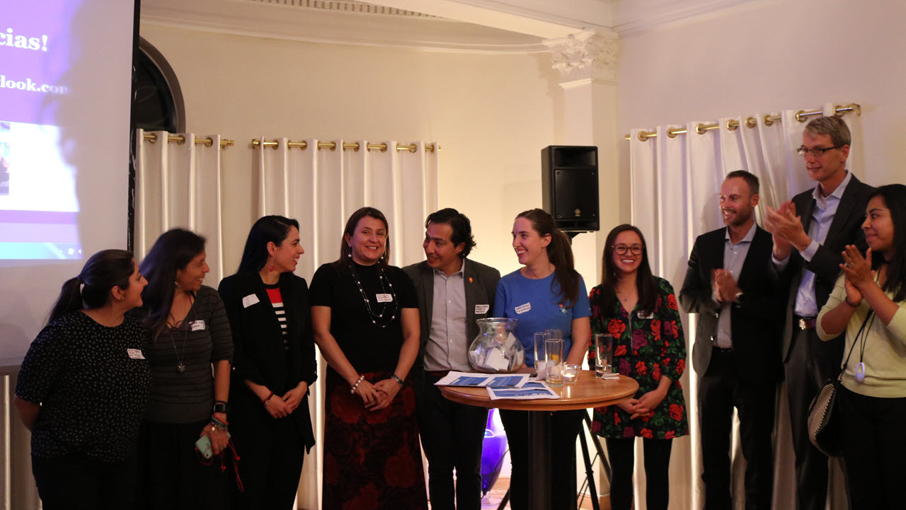 A group of Maria Recaman Koppel's colleagues stand together in a semi-circle, smiling and engaging with one another in a warmly lit room with white curtains and decorative molding. They are dressed in a mix of professional and casual attire, with some wearing name tags. A presentation screen is partially visible on the left side of the image with text in Spanish, and two people on the right are clapping, adding a celebratory feel to the scene.