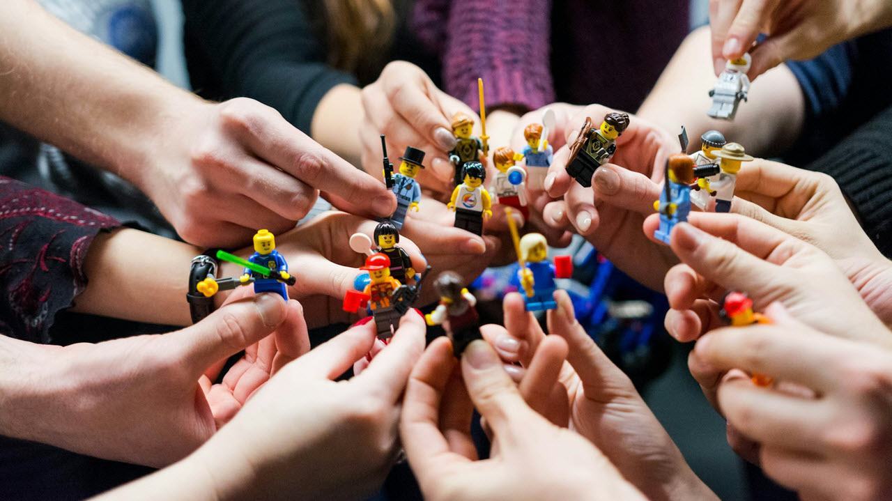 A group of people holding LEGO mini-figures in their hands, extending them toward the center of the frame. The mini-figures are diverse, depicting different characters and roles, such as a construction worker, a police officer, a ninja, and others.