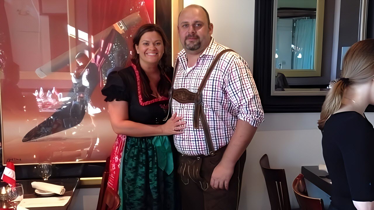 A smiling couple stands together indoors, dressed in traditional Bavarian attire. The woman is wearing a black dress with a red and green dirndl, while the man is wearing a checkered shirt with brown leather lederhosen.