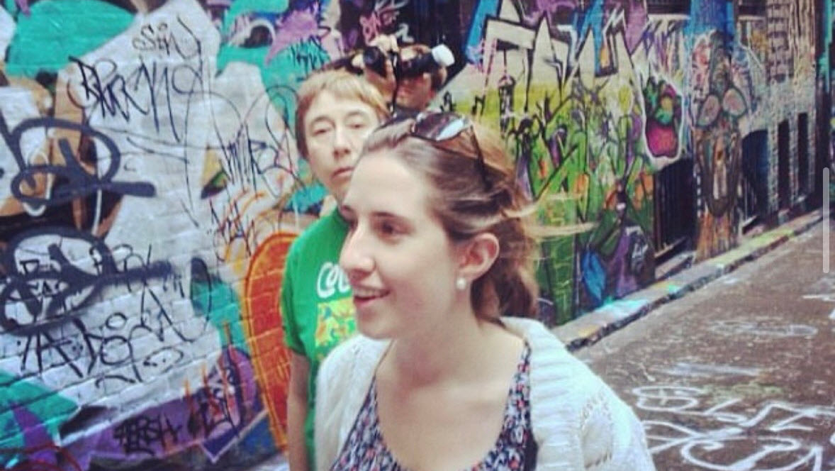 Maria Recaman walks through a vibrant, graffiti-covered alleyway in Melbourne with a smile on her face. She has her tied back, is wearing sunglasses on her head, a patterned top, and a light cardigan as she is in holiday mode. Behind her, a friend in a green shirt looks at the person taking the photo.